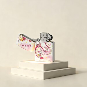 May include: A white metal flip-top lighter featuring a pink and yellow graphic of Hello Kitty characters and a long dragon-like creature. The design includes small stars and the text Hello Kitty in a cursive script on the open lid section.