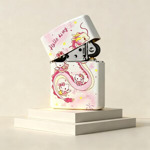 May include: A white metal flip-top lighter featuring a pink and yellow dragon graphic with Hello Kitty characters. The text Hello Kitty is printed in a cursive script on the lid. The lighter is open, revealing the internal metal mechanism.