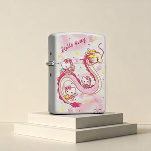 May include: A white metal flip-top lighter featuring a graphic of Hello Kitty characters and a long pink dragon. The design includes the text Hello Kitty in pink script, small yellow stars, and pink hearts against a soft pink background.