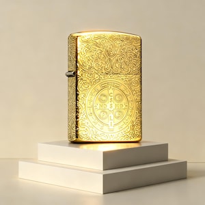 Puede incluir: Un encendedor de metal dorado con un intrincado patr&oacute;n de volutas en relieve en toda su superficie. La parte frontal muestra un dise&ntilde;o circular de la medalla de San Benito con las letras C S S M L N D S M D y V R S N S M V S M Q L I V B.
