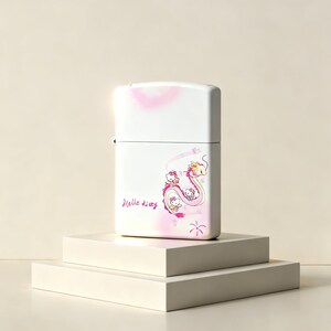 May include: A white rectangular metal lighter featuring a pink and yellow dragon graphic with small Hello Kitty characters. The text Hello Kitty is printed in pink script on the lower left side, with soft pink airbrushed accents on the surface.