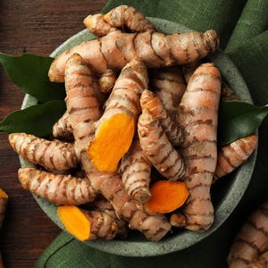 May include: A close-up shot of fresh turmeric roots in a gray bowl, with green leaves. The turmeric has a brown exterior and a vibrant orange interior. The bowl sits on a wooden surface with a green cloth.