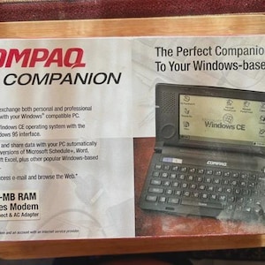 May include: A Compaq PC Companion box with a handheld computer featuring a screen displaying Windows CE. The box has the Compaq logo and text describing the device's features, including 2-MB RAM and a modem.