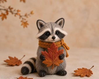 Pode incluir: Uma figura de guaxinim de lã feltrada com pelo cinzento e preto, usando um cachecol de malha amarelo-mostarda. O guaxinim segura uma grande folha de ácer texturizada em laranja e vermelho, com uma cauda às riscas e manchas pretas.