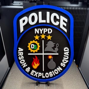 Puede incluir: Una placa de pared en forma de escudo con borde azul y centro negro. Presenta el texto blanco "POLICE" y "NYPD" sobre "ARSON & EXPLOSION SQUAD". El diseño incluye tres estrellas amarillas, un icono de engranaje verde "DB", una silueta de detective gris, una llama roja y una bomba gris.
