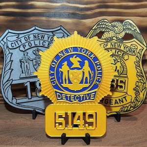 May include: Three decorative police badge replicas. The center gold badge features a blue circular emblem with the text City of New York Police Detective and the number 5149. Flanking it are a silver badge and a gold badge with an eagle emblem.