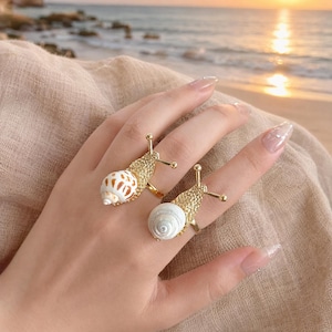 Puede incluir: Dos anillos dorados con diseño de caracol. Cada anillo tiene un cuerpo de metal dorado texturizado con dos antenas y una concha natural como caparazón. Una concha es blanca con manchas marrones y la otra es una espiral blanca lisa y uniforme.