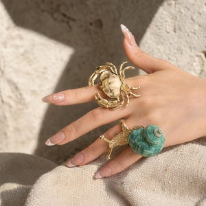 May include: A gold-toned ring shaped like a crab with a polished finish on the middle finger and a gold-toned ring featuring a textured body and a spiraled green shell shaped like a snail on the ring finger.