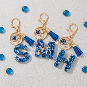 May include: Three gold-toned metal keychains featuring the letters S, M, and H. Each letter is crafted from blue resin with embedded gold flakes. Attached to each keychain is a blue tassel and a circular charm with a blue and white evil eye design.