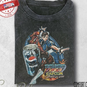 Puede incluir: Una camiseta gris carbón con efecto lavado al ácido que presenta un estampado gráfico del personaje Chun-Li de Tatsunoko vs. Capcom, junto a una lata de Pepsi. El texto en el gráfico dice Tatsunoko vs. Capcom Ultimate All-Stars.