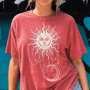 Puede incluir: Una camiseta de manga corta color rojo ladrillo con un estampado gráfico blanco de un sol con rostro humano, cadenas celestiales colgantes y una luna creciente. Se combina con pantalones cortos de mezclilla azul claro y gafas negras.