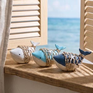 May include: Three whale figurines in white, light blue, and dark blue, each wrapped in a decorative tan jute fishing net with a small white starfish accent. The whales have smooth, rounded bodies and are positioned in a row on a wooden surface.