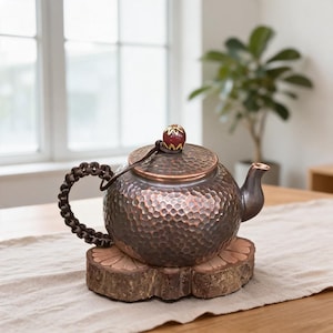 May include: A round, hammered copper teapot with a dark, weathered finish. It features a braided dark brown cord handle, a short spout, and a lid topped with a small, round, maroon-coloured knob accented with gold-coloured floral detailing.