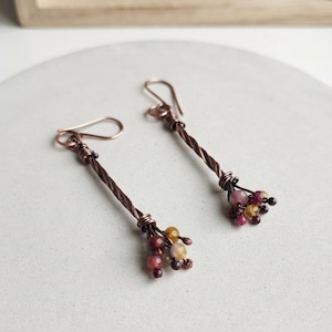 May include: A pair of dangle earrings featuring twisted copper wire stems that terminate in clusters of small, faceted, multicolored gemstone beads in shades of deep red, burgundy, and golden yellow, finished with simple copper ear wire hooks.