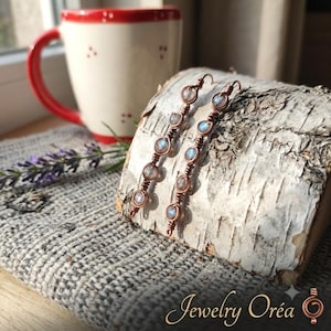 May include: A pair of long, dangling earrings crafted from copper wire wrapped around small, round, iridescent moonstone beads. Each earring features a vertical arrangement of four beads secured by intricate wire coils and a hook-style closure.