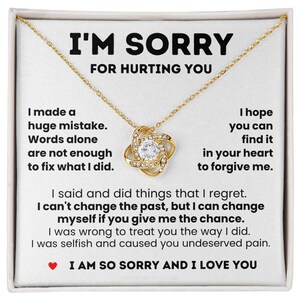 Op de afbeelding: Een goudkleurige ketting met een centrale knoop hanger met een heldere ronde steen, weergegeven op een witte kaart met zwarte tekst I'M SORRY FOR HURTING YOU, gevolgd door een verontschuldiging en een klein rood hartpictogram onderaan.