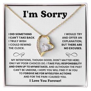 Peut inclure: Un collier doré avec un pendentif en forme de cœur orné d'un cristal rond central et de plus petits cristaux sur un côté. Le pendentif est présenté sur une carte portant le texte I'm Sorry et un long message sur la responsabilité.