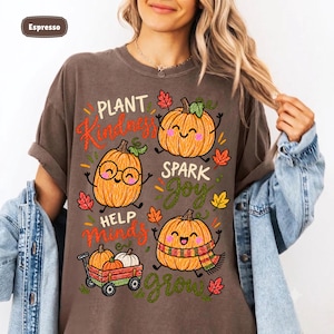 May include: An espresso-colored t-shirt featuring a graphic print of three smiling pumpkins and a small red wagon filled with pumpkins. The text reads "PLANT Kindness SPARK JOY HELP MINDS grow" in various colors, surrounded by small colorful autumn leaves.