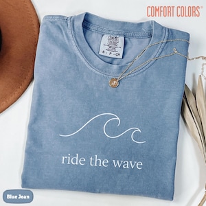 May include: A blue jean colored t-shirt featuring a white line drawing of an ocean wave and the text "ride the wave" printed below it. The shirt has a Comfort Colors brand label inside the collar and is paired with a gold chain necklace.