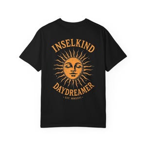 Könnte beinhalten: Ein schwarzes Kurzarm-T-Shirt mit einer großen orangefarbenen Grafik auf der Rückseite. Das Design zeigt eine Sonne mit einem menschlichen Gesicht, umgeben vom Text INSELKIND DAYDREAMER und EST. MMXXVI. Die Sonne hat Strahlen und kleine Sterne.