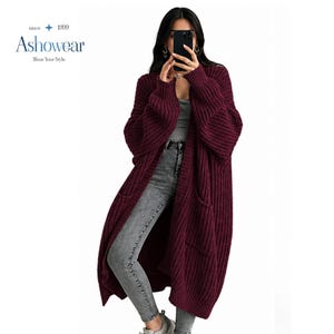 May include: A long, open-front burgundy cardigan made of a chunky ribbed knit fabric. The garment features long sleeves, two large front patch pockets, and a relaxed fit. The text "since 1999 Ashowear Wear Your Style" is visible in the top left corner.