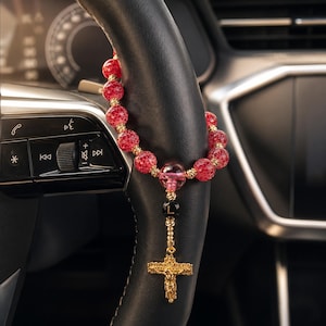 Pode incluir: Um rosário com contas de vidro craquelado vermelho, espaçadores dourados, uma conta rosa translúcida, uma conta preta com a letra L em dourado e um crucifixo dourado. O rosário está pendurado num volante de couro preto.