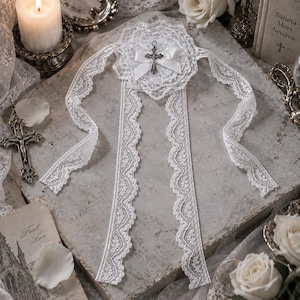 May include: A white lace accessory featuring a central circular lace rosette adorned with a small white satin ribbon bow and a silver-toned metal crucifix pendant. Three long, scalloped lace ribbons extend from the central rosette.