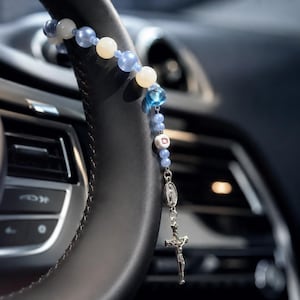 Op de afbeelding: Een rozenkrans met lichtblauwe, witte en doorschijnend blauwe kralen, een witte kubuskraal met de letter D, een zilverkleurige metalen medaille en een zilverkleurig kruisbeeld. De rozenkrans hangt over een zwart met leer bekleed stuurwiel.