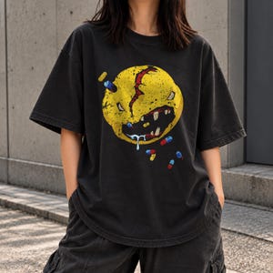 Puede incluir: Una camiseta gris carbón de manga corta con un gran gráfico circular amarillo desgastado. El diseño muestra un rostro monstruoso agrietado con dientes afilados, baba y varias cápsulas de pastillas pequeñas y coloridas dispersas.