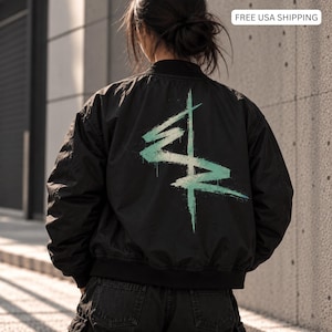 May include: A black bomber jacket featuring a large, light green, brushstroke-style graphic design on the back. The jacket has a ribbed collar, long sleeves, and a solid black finish. The design consists of intersecting lines with a textured, paint-like appearance.