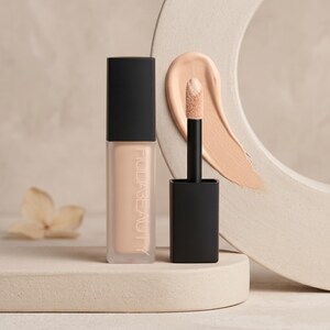 Puede incluir: Un corrector l&iacute;quido Huda Beauty en un frasco rectangular de vidrio esmerilado con tapa negra, acompa&ntilde;ado de un aplicador negro con una muestra de corrector beige en la punta, mostrando el nombre de la marca verticalmente.