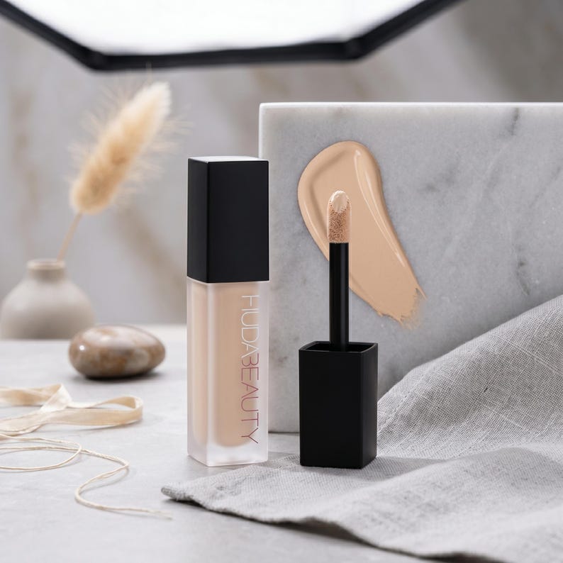 Puede incluir: Un frasco cuadrado de vidrio de corrector l&iacute;quido Huda Beauty con tapa negra y aplicador a juego. El nombre de la marca est&aacute; impreso verticalmente en blanco. Se observa una muestra de corrector beige sobre un bloque de m&aacute;rmol.