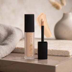 Puede incluir: Un corrector l&iacute;quido Huda Beauty en un frasco rectangular de vidrio esmerilado con tapa negra, acompa&ntilde;ado de un aplicador negro con una muestra de corrector beige. El nombre de la marca Huda Beauty est&aacute; impreso verticalmente.