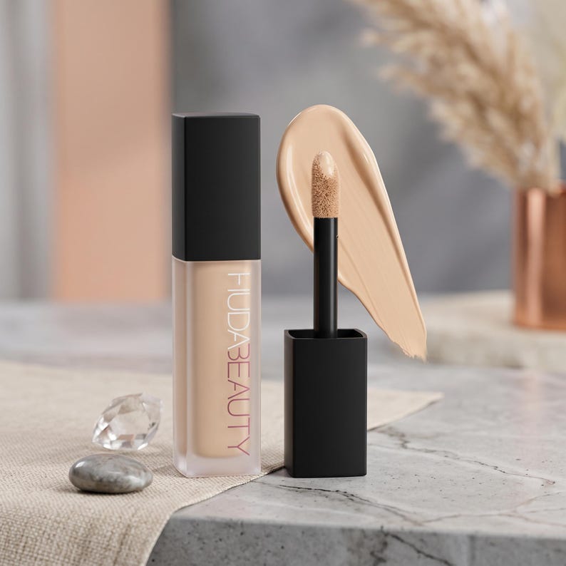Puede incluir: Un corrector l&iacute;quido Huda Beauty en un frasco rectangular transparente con tapa negra. El aplicador est&aacute; fuera, mostrando una muestra de corrector beige en la punta. El nombre de la marca Huda Beauty est&aacute; impreso verticalmente en negro.