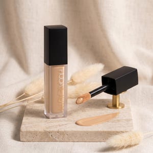 Puede incluir: Un corrector l&iacute;quido Huda Beauty en un frasco de vidrio rectangular transparente con tapa negra. Al lado, un aplicador negro con punta de esponja beige contiene un poco de producto y se observa una muestra suave del l&iacute;quido beige.