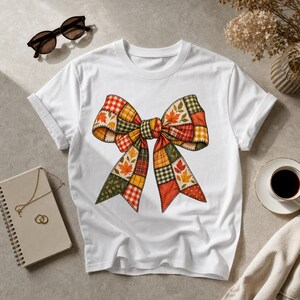 Op de afbeelding: Een wit t-shirt met korte mouwen en een grote centrale afbeelding van een strik. De strik is samengesteld uit een patchworkpatroon in tinten oranje, rood, geel en groen, versierd met diverse herfstbladmotieven en ruitpatronen.
