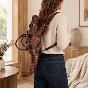 Op de afbeelding: Een donkerbruine leren rugzak in de vorm van een krokodil met een textuur van schubben. De tas heeft een lange staart, vier poten en een gedetailleerde kop. Het bevat donkerbruine leren schouderbanden en twee handvatten om te dragen.