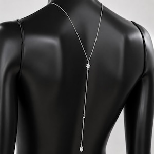 K&ouml;nnte beinhalten: Eine silberfarbene R&uuml;ckenkette mit einer zarten Kette, die den R&uuml;cken hinunterf&auml;llt. Das Design umfasst einen zentralen sternf&ouml;rmigen Kristallakzent, eine kleine runde Perle und einen einzelnen tropfenf&ouml;rmigen Kristallanh&auml;nger am unteren Ende.