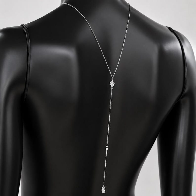 K&ouml;nnte beinhalten: Eine silberfarbene R&uuml;ckenkette mit einer zarten Kette, die den R&uuml;cken hinunterf&auml;llt. Das Design umfasst einen zentralen sternf&ouml;rmigen Kristallakzent, eine kleine runde Perle und einen einzelnen tropfenf&ouml;rmigen Kristallanh&auml;nger am unteren Ende.