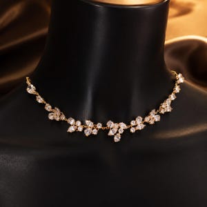 K&ouml;nnte beinhalten: Eine goldfarbene Halskette mit einer Reihe von klaren, birnenf&ouml;rmigen und rund geschliffenen Kristallen, die in einem zarten, rankenartigen Muster angeordnet sind. Die Kristalle sind durch eine d&uuml;nne goldene Kette verbunden.