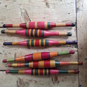 May include: Six cylindrical wooden spindles arranged horizontally, each featuring a unique pattern of painted bands in red, green, yellow, orange, and purple. The spindles have tapered ends and rounded tips, showcasing a smooth, polished finish.