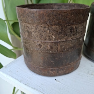 May include: A cylindrical metal container with a heavily rusted, dark brown patina. The exterior features horizontal metal bands reinforced with visible rivets, creating a rugged, weathered texture across the entire surface of the vessel.