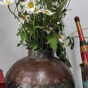 May include: A large, spherical, riveted metal vessel with a dark, weathered patina, filled with a bouquet of white daisies with yellow centres. Beside it stands a tall, slender, multi-coloured wooden tool and a small, bright red, lidded container.