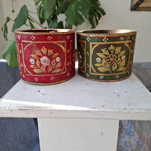 May include: Two cylindrical wooden containers featuring hand-painted floral motifs. One container is red with gold-toned borders and white dot accents, while the other is dark green with similar gold-toned borders and white dot detailing.