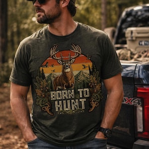 Peut inclure: Un t-shirt à manches courtes vert olive avec une impression graphique représentant un cerf aux grands bois au centre d'un paysage de montagne au coucher du soleil. Le texte BORN TO HUNT est imprimé sous le cerf, entouré de feuilles de chêne.