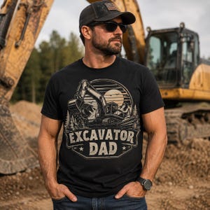 Puede incluir: Una camiseta negra de manga corta con un estampado gráfico de una excavadora frente a un paisaje de sol y montañas. El texto EXCAVATOR DAD está impreso en una fuente negrita y desgastada debajo de la ilustración, acompañado por dos pequeñas estrellas.