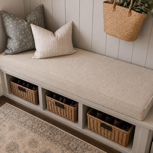 May include: A light gray entryway bench featuring a thick beige fabric cushion, three open storage compartments with woven wicker baskets, and two decorative throw pillows in gray floral and beige striped patterns with a hanging straw bag.