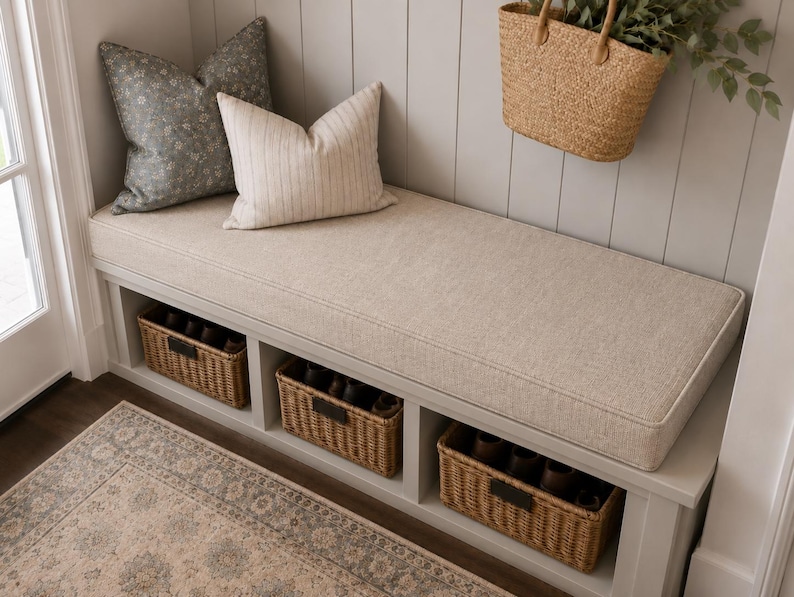 May include: A light gray entryway bench featuring a thick beige fabric cushion, three open storage compartments with woven wicker baskets, and two decorative throw pillows in gray floral and beige striped patterns with a hanging straw bag.