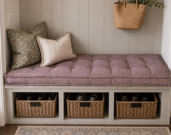 May include: A light gray entryway bench featuring a tufted mauve cushion, two decorative pillows, and three woven storage baskets. One pillow is olive green with a white floral pattern, and the other is cream with thin vertical stripes.