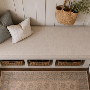 May include: A white entryway bench with a beige upholstered seat cushion, featuring three woven storage baskets underneath. Two decorative pillows, one solid gray and one white with thin stripes, sit on the bench alongside a hanging straw tote bag.